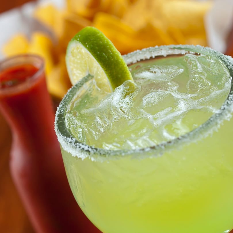Close-up of a Padrino's margarita with a lime wedge, salt rim, and ice, with salsa and chips in the background.