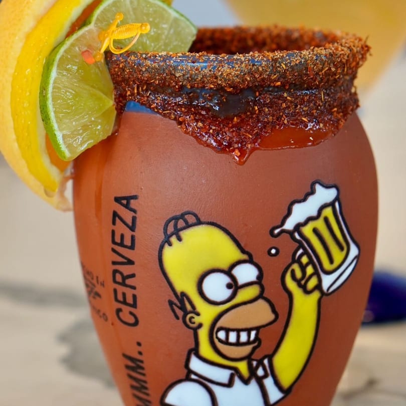 A Padrino's michelada in a Homer Simpson mug, garnished with lime and lemon slices.