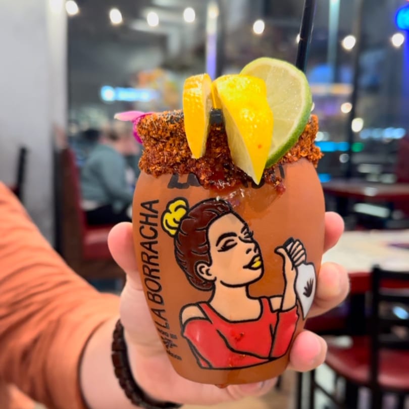 A hand holds a Padrino's michelada in a clay cup with a woman's illustration, garnished with lime and lemon.