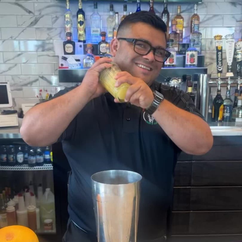 A smiling Padrino's bartender in glasses shakes a yellow cocktail over a metal shaker.