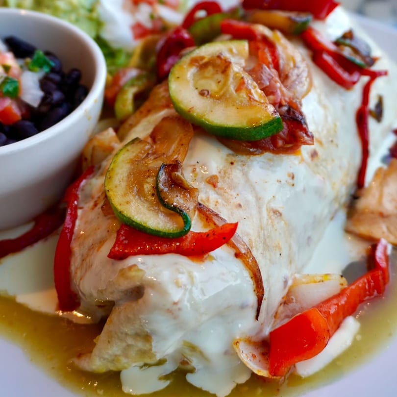 Padrino's Mexican burrito covered in white sauce, red peppers, and zucchini, with a side of black beans.