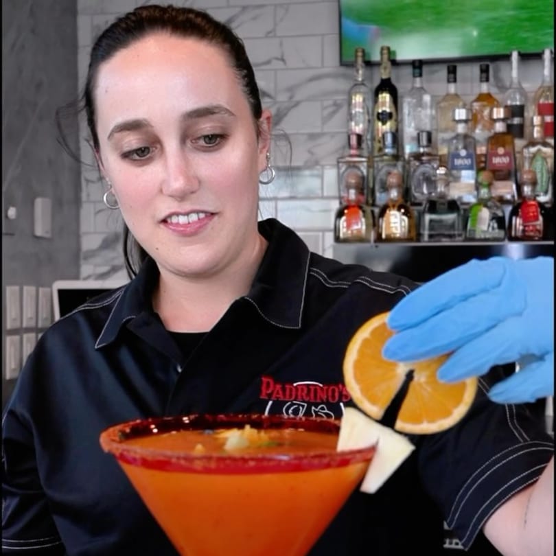 A Padrino's bartender garnishes a vibrant orange cocktail with an orange slice and pineapple.