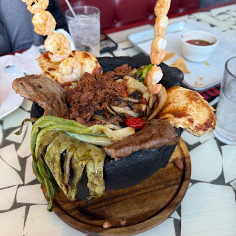 A large molcajete filled with grilled meats, shrimp skewers, vegetables, and tortillas on a wooden tray.