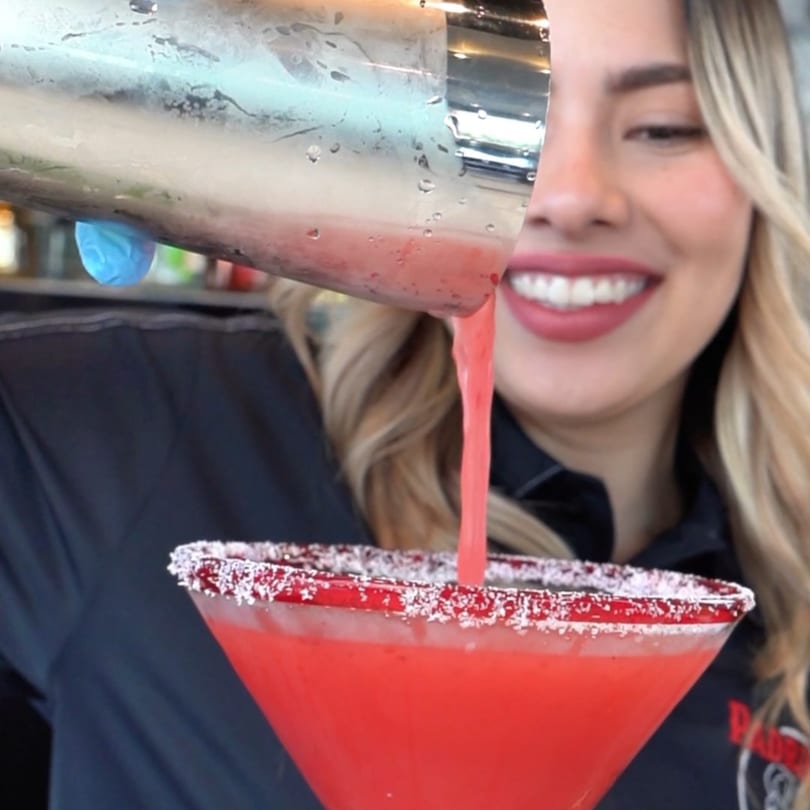 A smiling Padrino's staff member pours a red margarita from a shaker into a salt-rimmed glass.