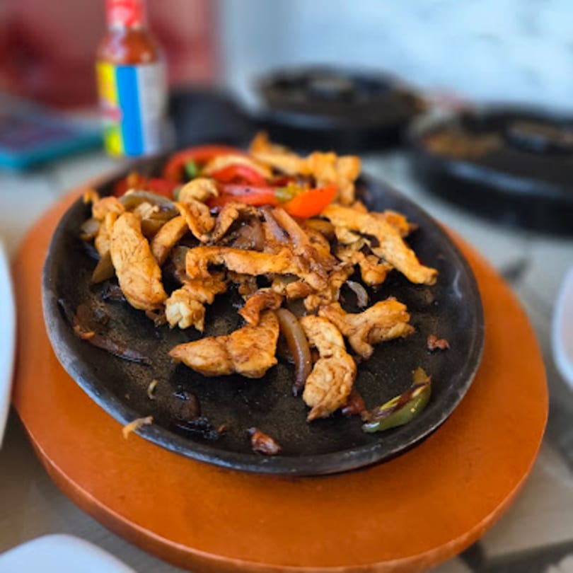 Sizzling chicken fajitas with onions and peppers on a cast iron skillet, with a hot sauce bottle in the background.