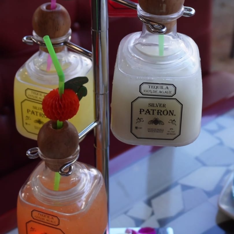 Three mini Patron tequila bottles, filled with colorful cocktails, hanging from a metal stand.