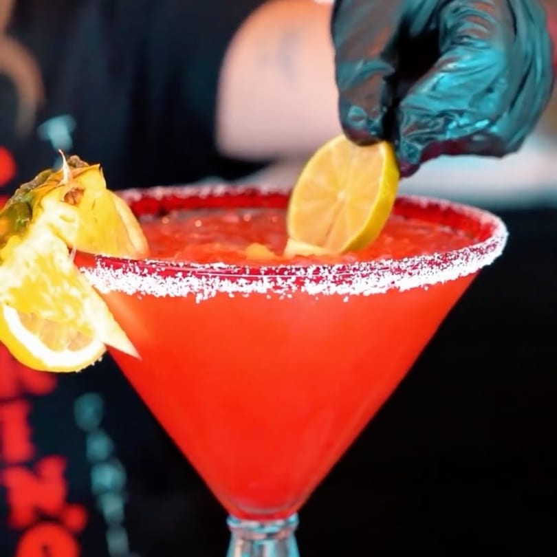 A hand in a black glove garnishes a red margarita with a lime wedge and pineapple.