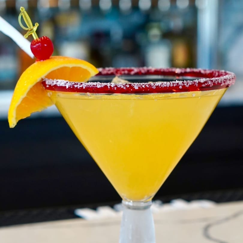 A vibrant yellow Padrino's margarita in a martini glass, garnished with an orange slice and cherry.