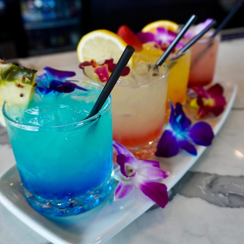 A tray of colorful Padrino's cocktails, garnished with fruit slices and vibrant orchids.